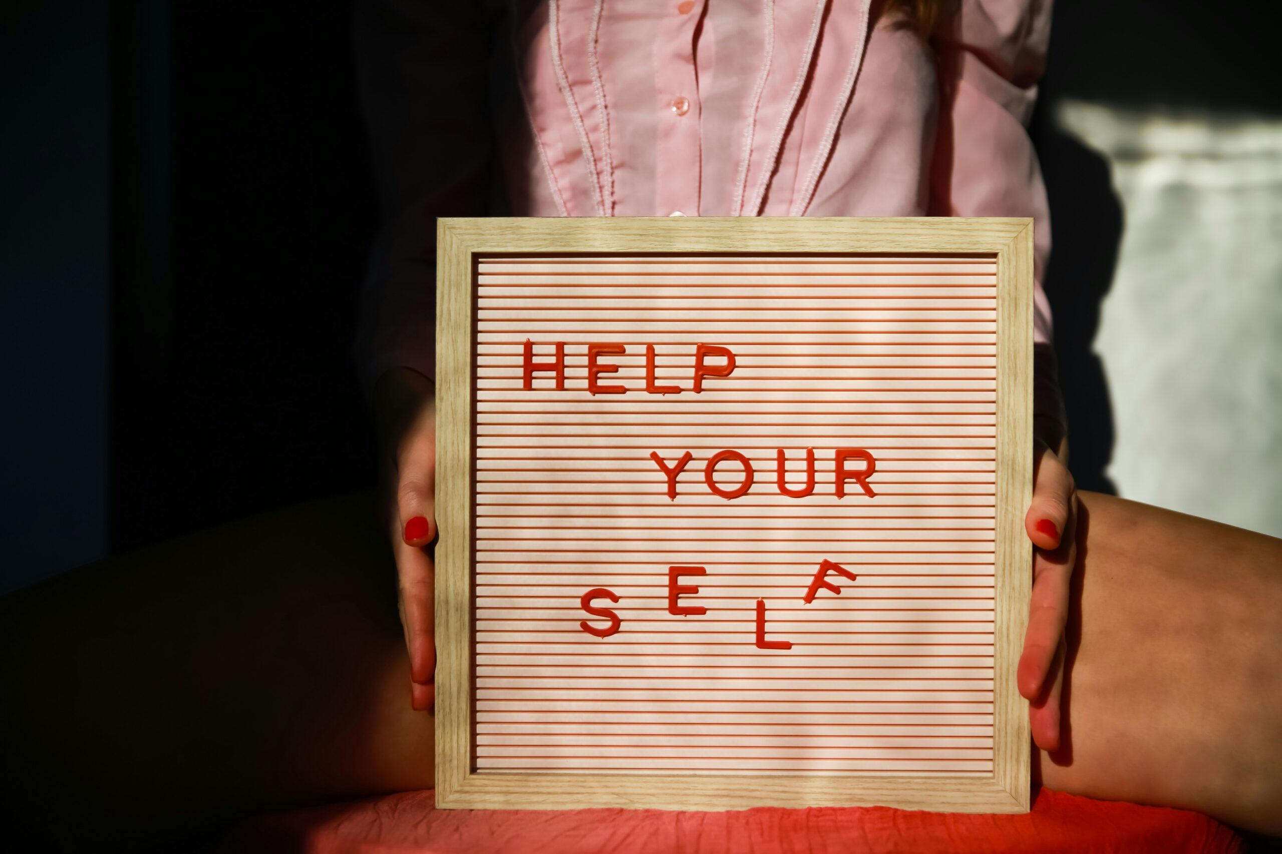 Seseorang memegang papan dengan tulisan "HELP YOUR SELF" yang sebagian hurufnya terjatuh.