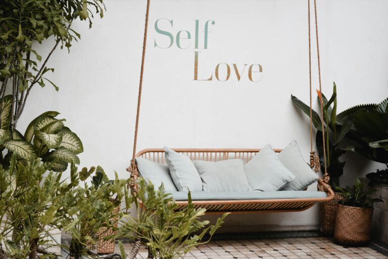 Sebuah bangku ayunan rotan dengan bantal berwarna hijau pastel di taman yang asri, dengan tulisan "Self Love" di dinding.