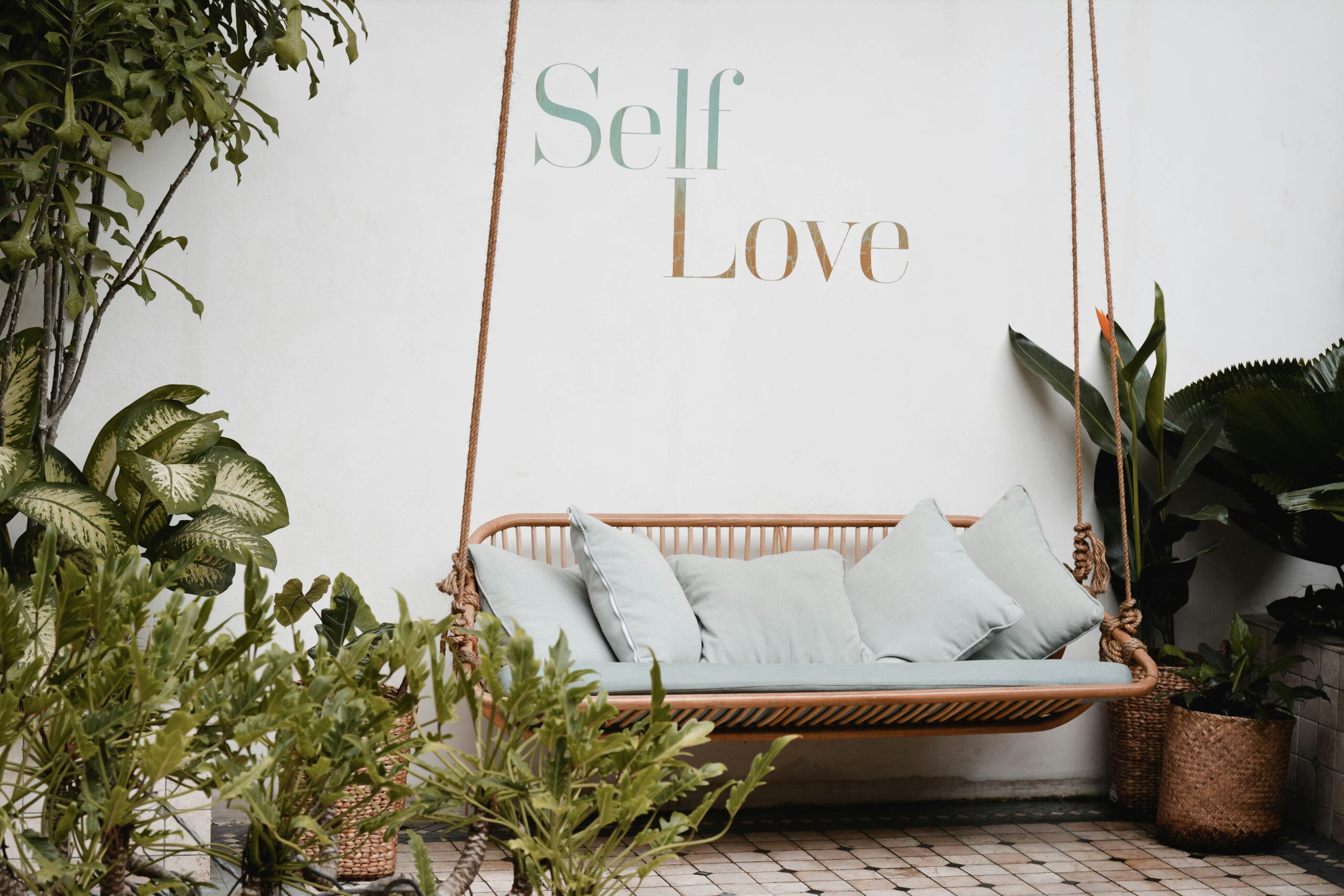 Sebuah bangku ayunan rotan dengan bantal berwarna hijau pastel di taman yang asri, dengan tulisan "Self Love" di dinding.