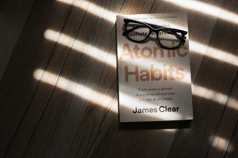 Buku Atomic Habits karya James Clear dengan kacamata di atasnya, diterangi cahaya matahari melalui jendela.