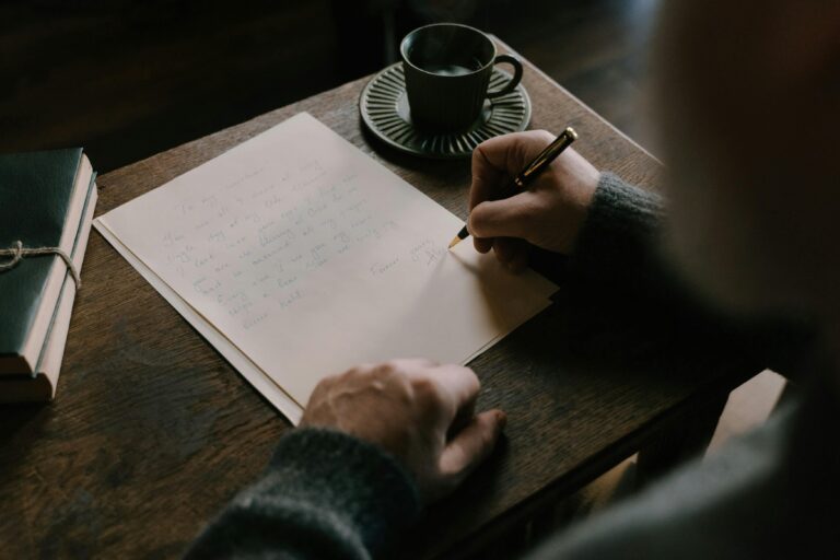 Seorang pria sedang menulis surat di atas meja kayu dengan secangkir kopi di sampingnya.