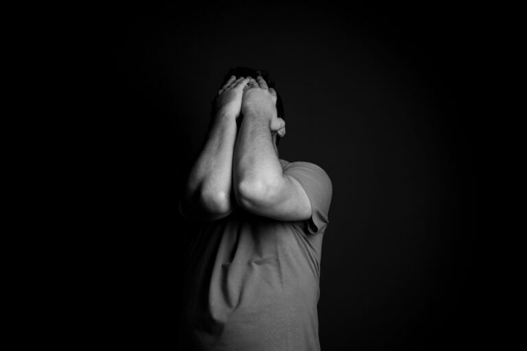 Seorang pria menutupi wajahnya dengan kedua tangan dalam suasana gelap, mencerminkan tekanan emosional dan kecemasan.